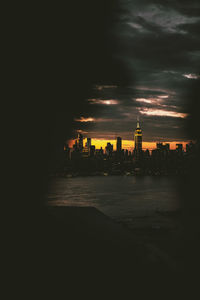 Illuminated buildings in city by river against sky during sunset