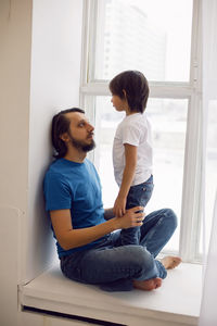 Father in a blue t-shirt and a son in a white t-shirt. sitting on the windowsill of a large window