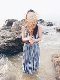 Woman covering face by flowers at beach against sky