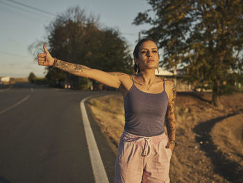 Serious tattooed female hitchhiker standing on road and hitchhiking with outstretched arm in summer at sunset