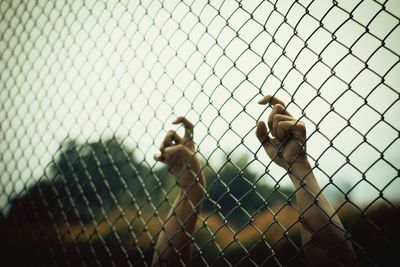 Cropped hands of person holding chainlink fence against sky