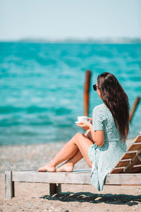 Rear view of woman holding coffee cup sitting at beach