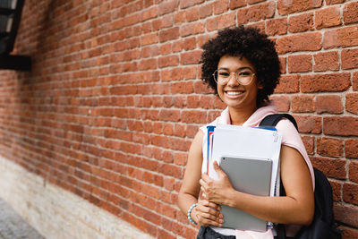 Portrait of smiling young woman using phone against brick wall
