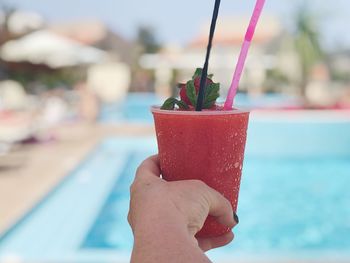 Close-up of hand holding an cold strawberry drink with drinking straws in front of a swimming pool 