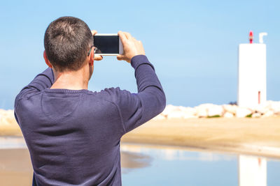 Rear view of man photographing with smart phone against sky during sunny day