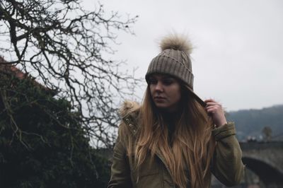 Young woman wearing knit hat while walking by bare trees against sky