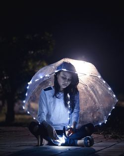 Young woman holding umbrella while sitting at night