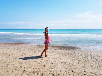 Woman holding champagne bottle standing on beach against clear blue sky