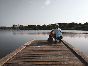 Rear view of woman sitting on pier over lake against sky