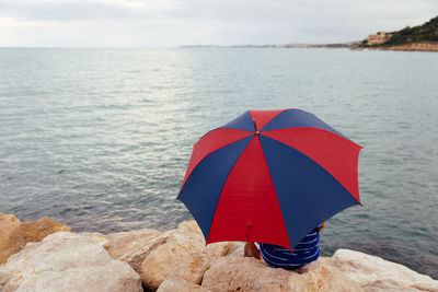 From behind anonymous man sitting on the rocks with an umbrella by the sea while it rains