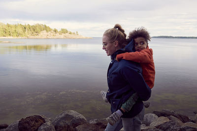 Side view of young mother giving piggyback to son at lakeshore