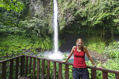 Portrait of happy woman standing against waterfall at forest