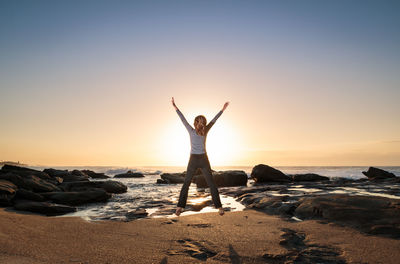 Rear view of excited woman jumping over beach against sky during sunrise