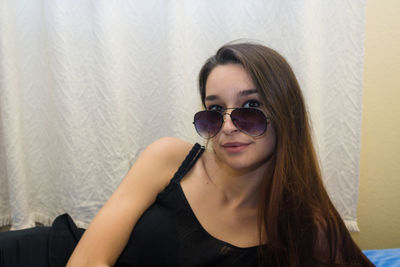 Portrait of young woman with long hair wearing sunglasses against curtain at home