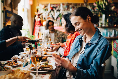 Smiling young woman using smart phone while sitting with friends at table in restaurant during dinner party