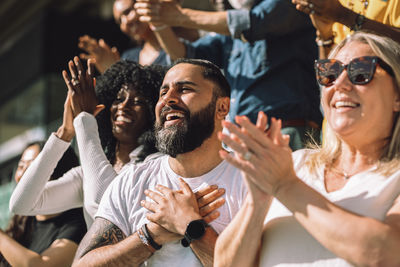 Happy bearded man celebrating with hands on chest surrounded by fans in stadium stand on sunny day