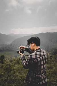 Young man photographing while standing on mountain against sky