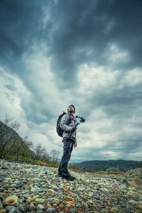 Low angle view of hiker holding camera while standing on field against cloudy sky