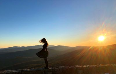 Side view of woman standing on stone wall by mountains at sunset