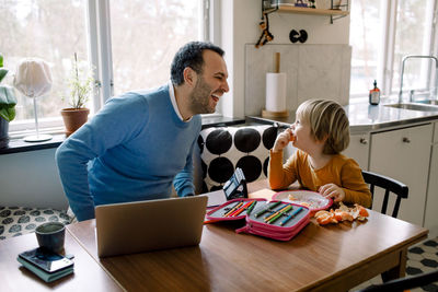 Playful father looking at daughter while using laptop at home