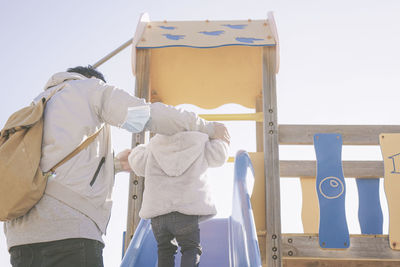 Rear view of man helping daughter to walk on slide at playground