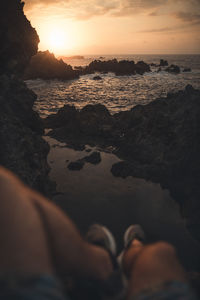 Low section of person on rocks at beach against sky during sunset