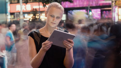 Portrait of woman holding smart phone while standing on illuminated laptop