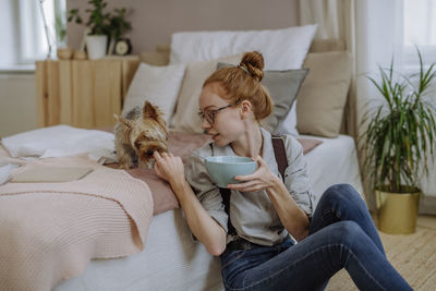 Woman wearing eyeglasses feeding dog sitting on bed at home