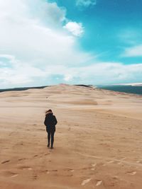 Rear view of young woman in warm clothing walking at beach against cloudy sky