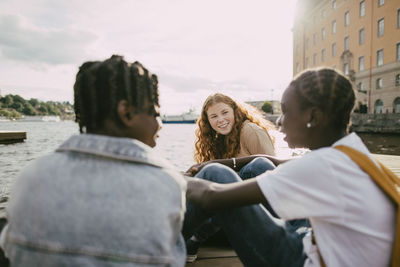 Smiling teenage girl talking with male friends near lake at sunny day