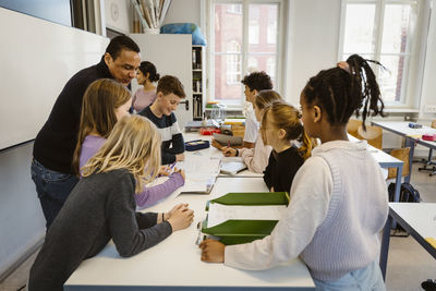 Male and female students studying with teacher at desk in classroom