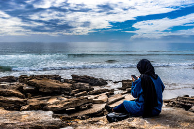 Rear view of woman sitting on rock by sea against sky