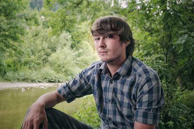 Portrait of handsome young man sitting at lakeshore against plants in forest