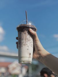 Cropped hand holding iced coffee against sky