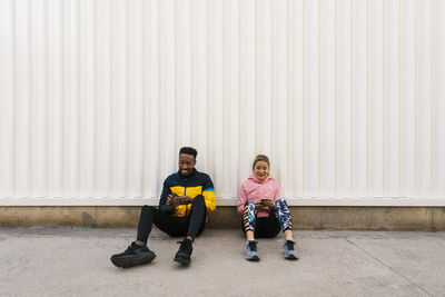 Smiling male and female athlete using mobile phone while sitting on floor against wall