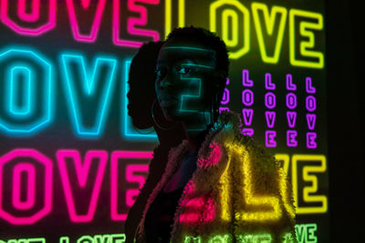 African american woman in trendy fur coat looking at camera while standing near dark wall with bright neon love inscriptions