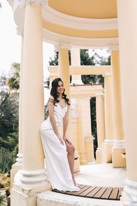 A beautiful brunette lady in an elegant wedding dress poses among the columns in the old city park