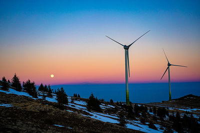 Wind turbines on snow covered landscape against sky during sunset