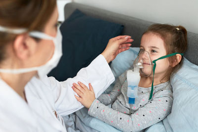 High angle view of female doctor wearing mask examining girl sitting on bed