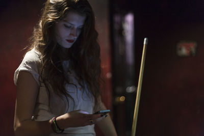 Young woman using mobile phone while holding pool cue