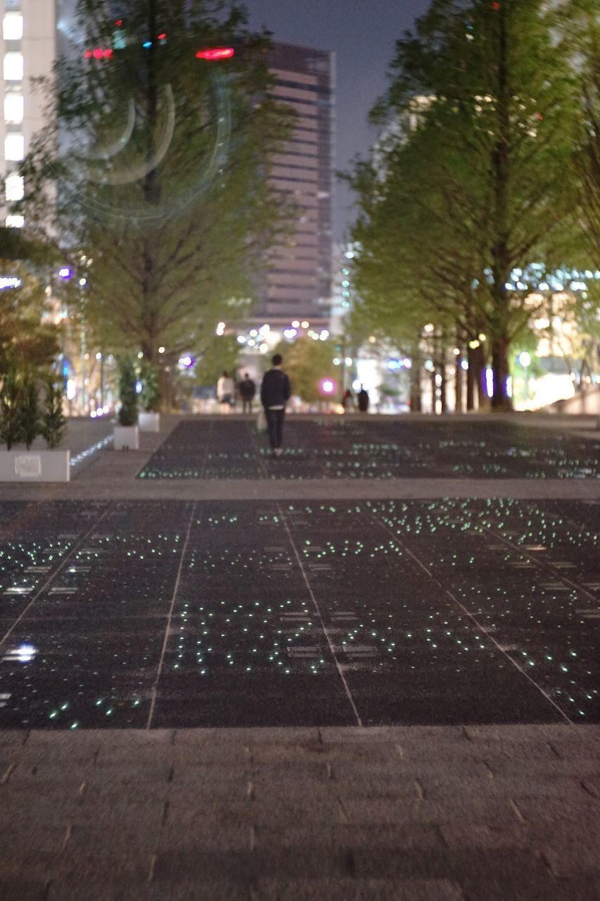 MAN WALKING ON FOOTPATH BY ILLUMINATED CITY