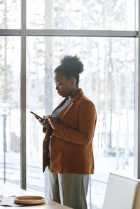 Young businesswoman reading news on smart phone standing by glass window in office