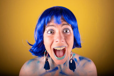 Close-up portrait of young female model with blue wig shouting against yellow background