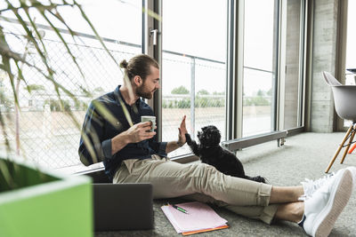 Young businessman with laptop sitting on the floor in office playing with dog