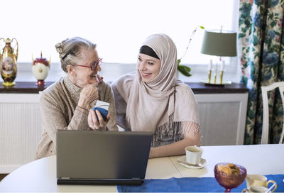 Happy young female home caregiver with senior woman doing bank transaction on laptop
