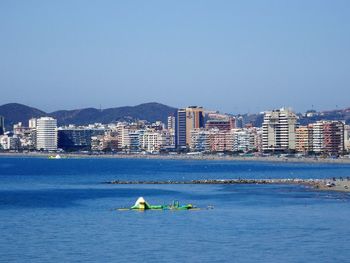 People in sea by cityscape against clear blue sky