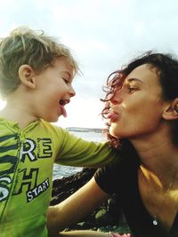 Woman sticking out tongue with son against sky