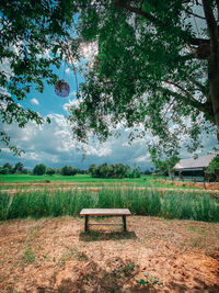 Bench on field by trees against sky