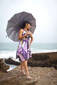 Full length of mature woman with umbrella standing on beach against sky