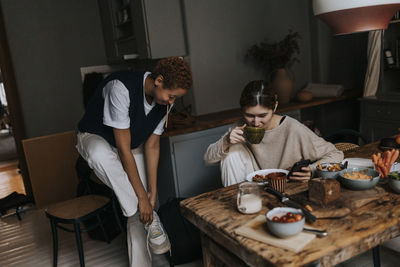 Non-binary person drinking coffee and using smart phone by friend wearing shoes at home